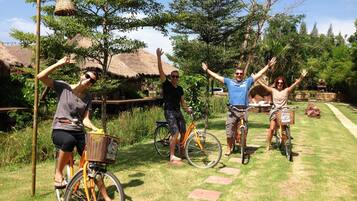 A group of people on bicycles in a garden with a thatched-roof structure and a bamboo pole.