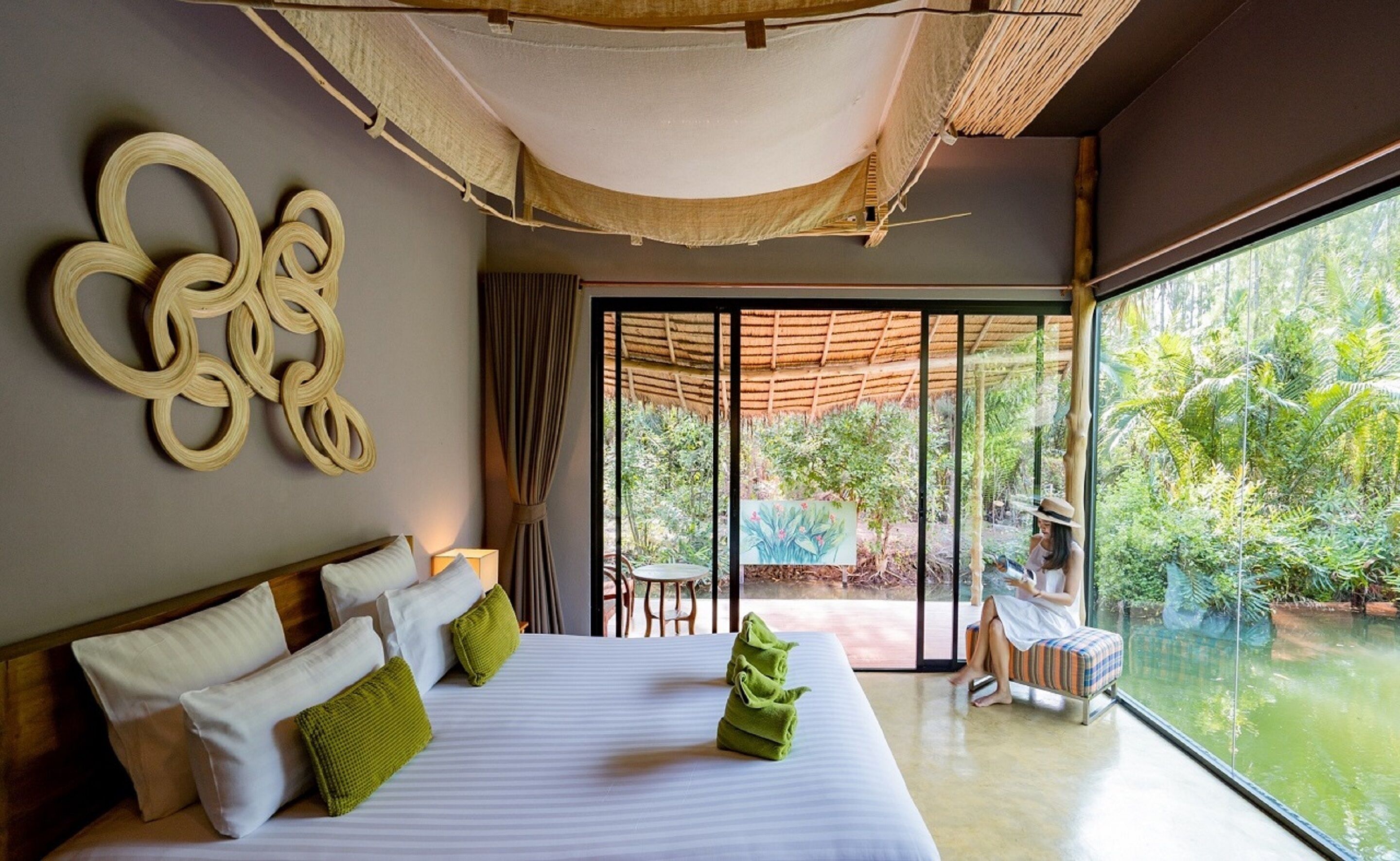 Una habitación de hotel moderna con una cama grande, vista a un jardín exuberante a través de una puerta corrediza de vidrio y una pieza decorativa original en la pared.