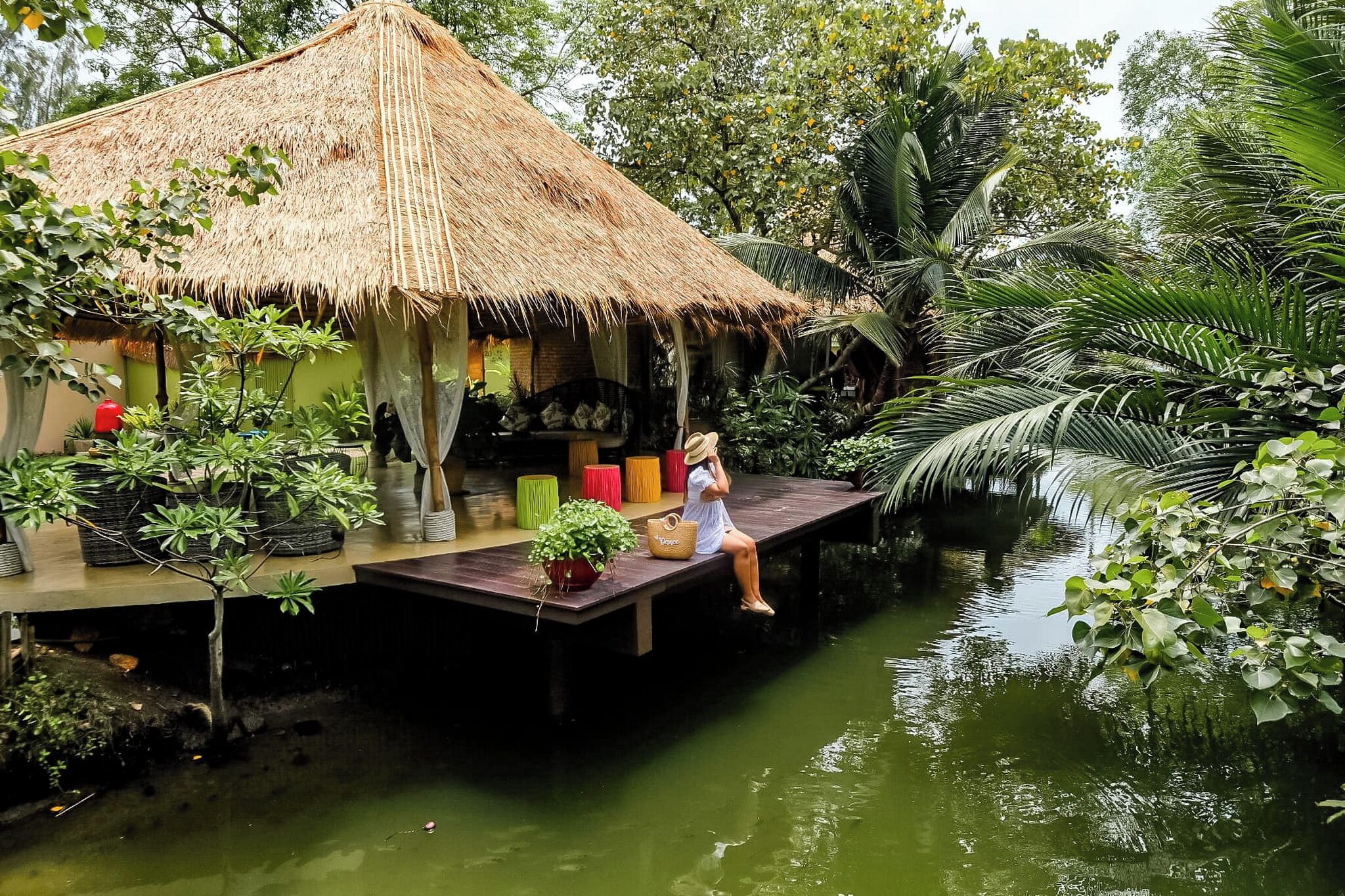 A thatched-roof structure with a wooden platform extending over water, surrounded by lush greenery.