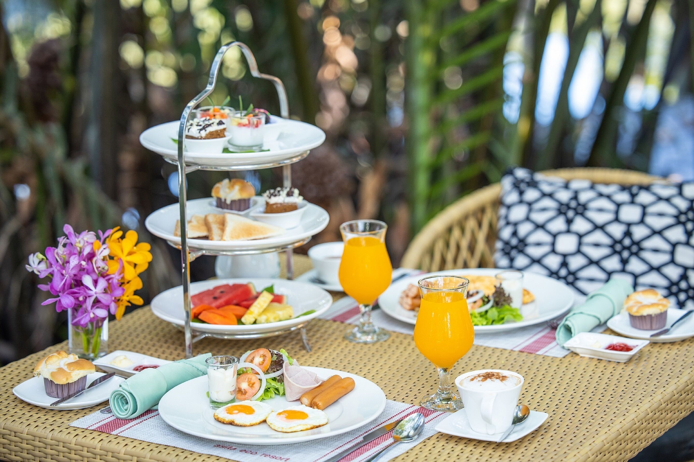Un présentoir à trois niveaux garni de diverses pâtisseries, une table en osier avec des assiettes de nourriture, un verre de jus d’orange et un arrangement floral.