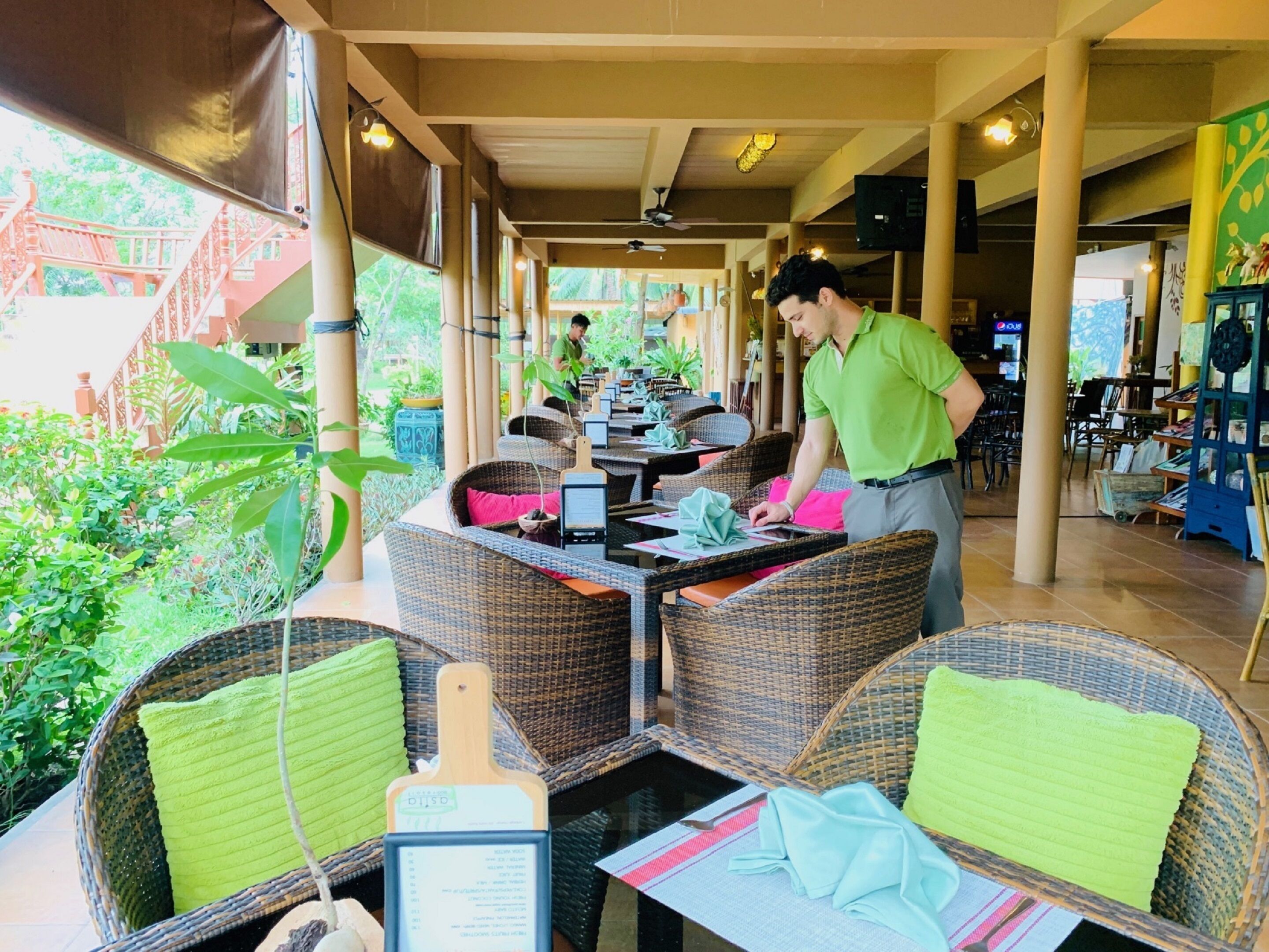 A restaurant with wicker furniture and green cushions, a waiter attending to a table, and a view of greenery outside.