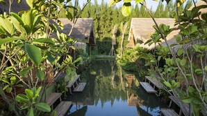 Un área de piscina tranquila al aire libre con bancos de madera y estructuras de techo de paja, rodeada de exuberante vegetación.