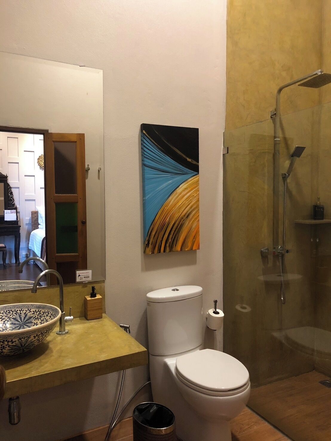 A bathroom with a glass shower enclosure, a white toilet, a sink with a decorative bowl, and a painting on the wall.