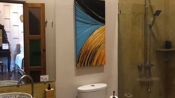 A bathroom with a glass shower enclosure, a white toilet, a sink with a decorative bowl, and a painting on the wall.