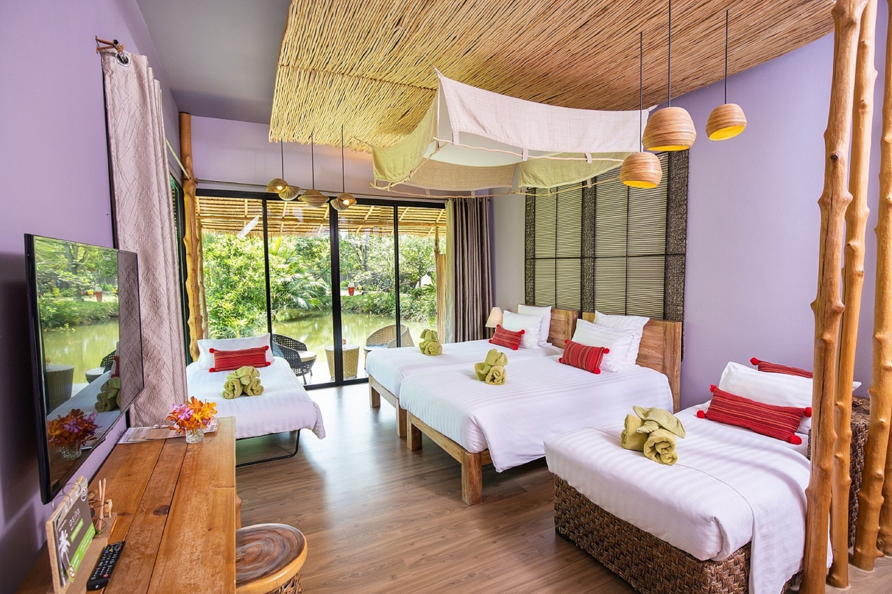 A hotel room with two beds, a wooden desk, a TV, and large windows with a view of greenery.