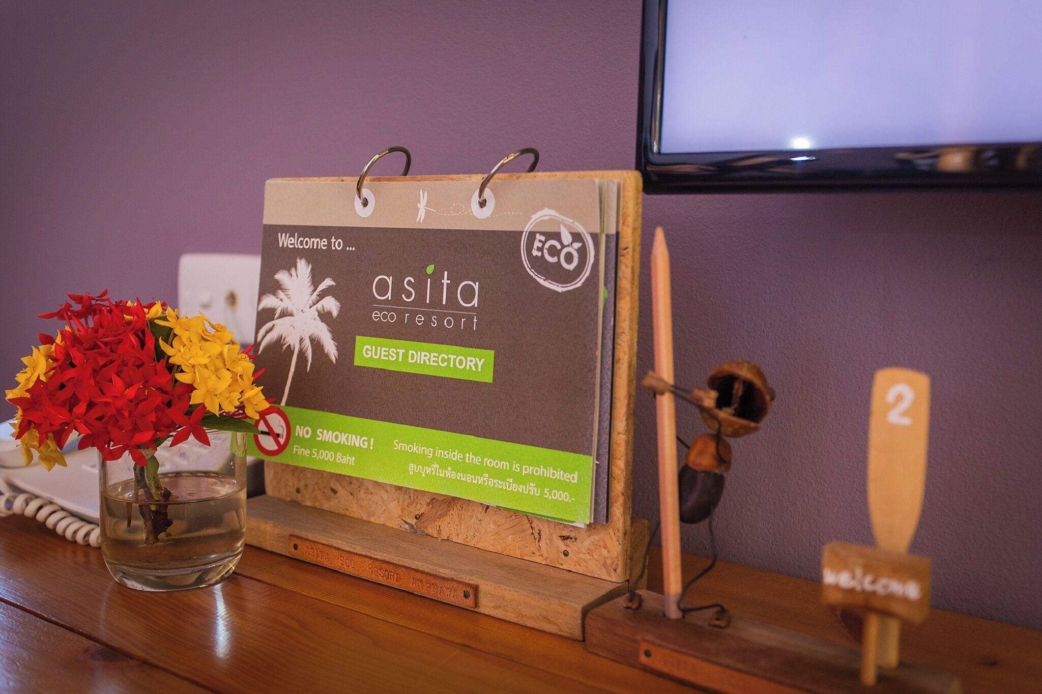A wooden guest directory at Asita eco resort with a 'NO SMOKING' sign, a small vase with flowers, and a wooden welcome sign.