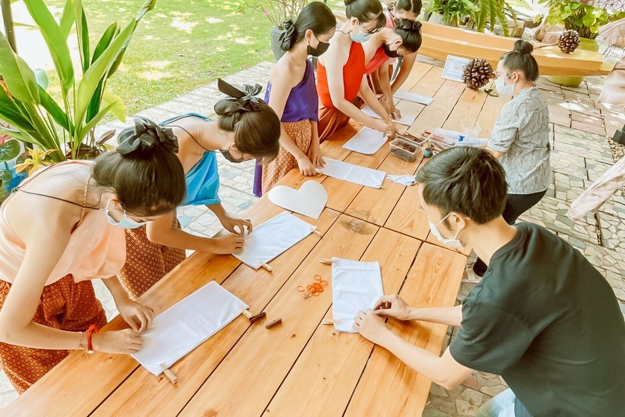 A group of people seated around a wooden table outdoors, engaged in a craft activity.