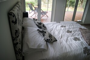 A bed with white bedding and black floral pillows, a headboard with a black and white pattern, and a view of an outdoor deck with chairs. - Eagles Nest (Merrijig)