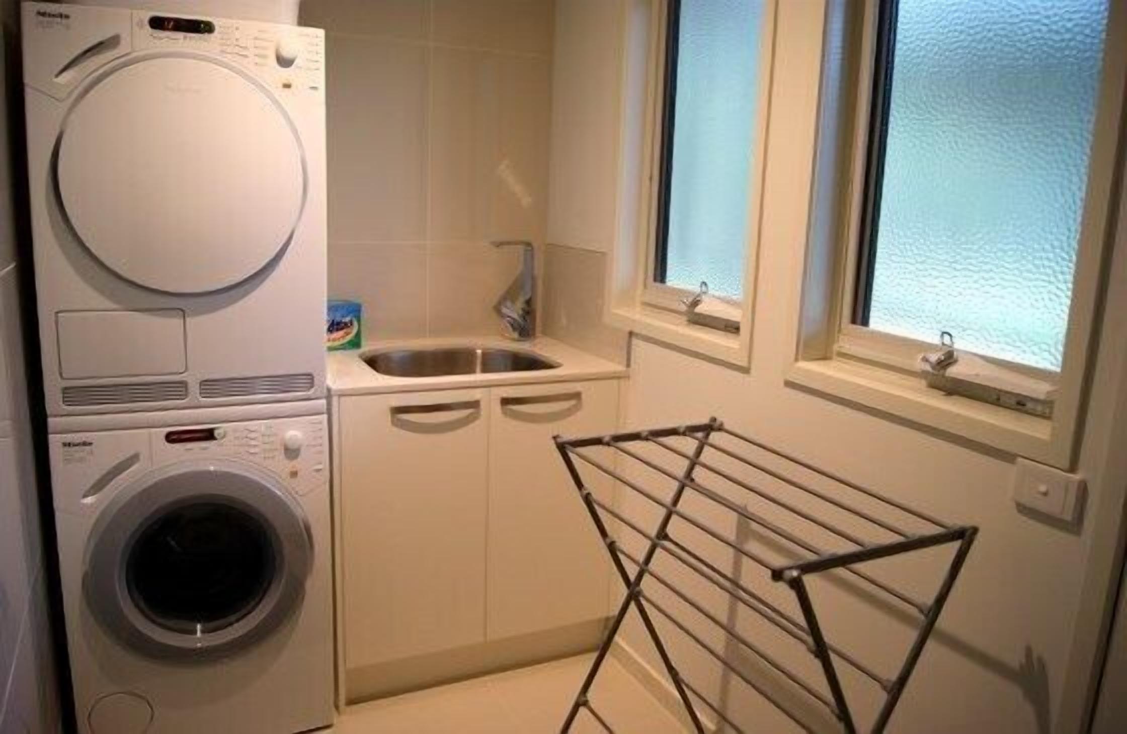 A compact laundry room with a front-loading washer and dryer, a sink, and a drying rack.