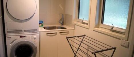 A compact laundry room with a front-loading washer and dryer, a sink, and a drying rack.