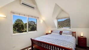 A bedroom with a sloped ceiling, a large bed with a wooden frame, two bedside tables with lamps, and a view of a forest through a window. - Jarrah Lodge (Merrijig)