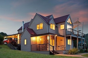 A two-story house with a covered porch, a stone fireplace, and a car parked in the driveway. - Jarrah Lodge (Merrijig)