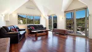 A spacious living room with a high ceiling, a black leather sofa, a wooden coffee table, and a sliding glass door leading to an outdoor area. - Jarrah Lodge (Merrijig)