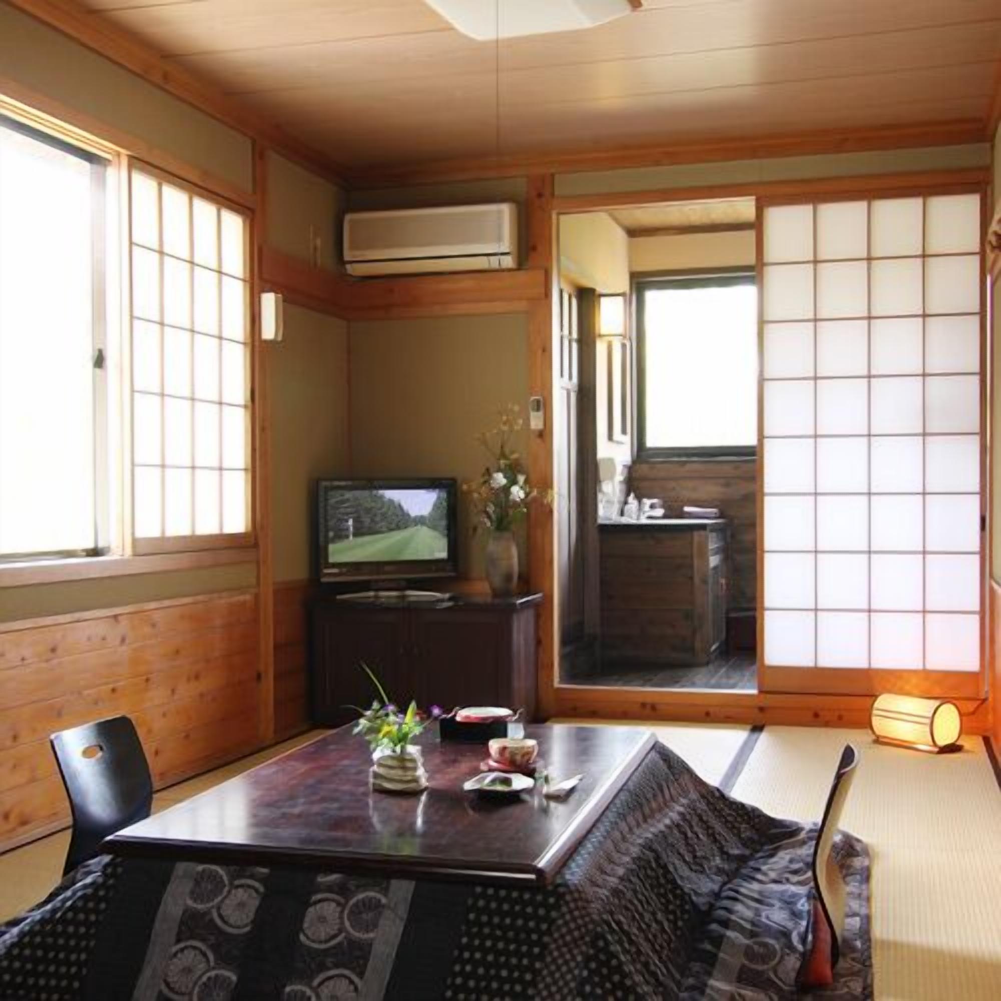 Una habitación japonesa tradicional con suelo de tatami, una mesa baja con un florero y flores, un televisor y puertas correderas que dan a otra habitación.