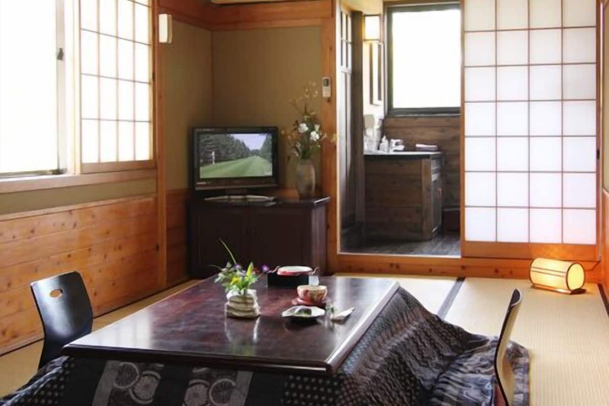 Um quarto japonês tradicional com tatames, uma mesa baixa com um vaso e flores, uma televisão e portas deslizantes que dão para outro cômodo.