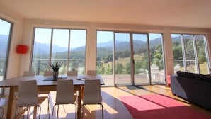 A spacious room with a large dining table, several chairs, and a sofa. The room has large windows offering a view of the countryside and mountains.