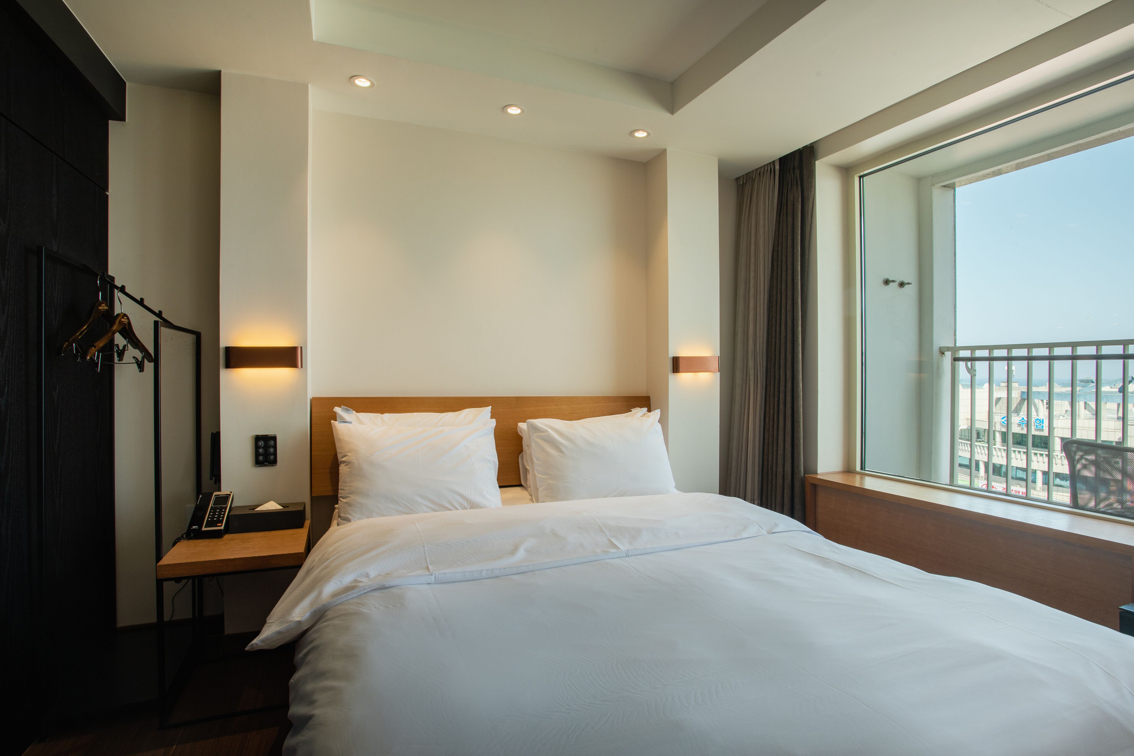 A hotel room with a large bed, a bedside table with a phone, and a window with a view of the city.