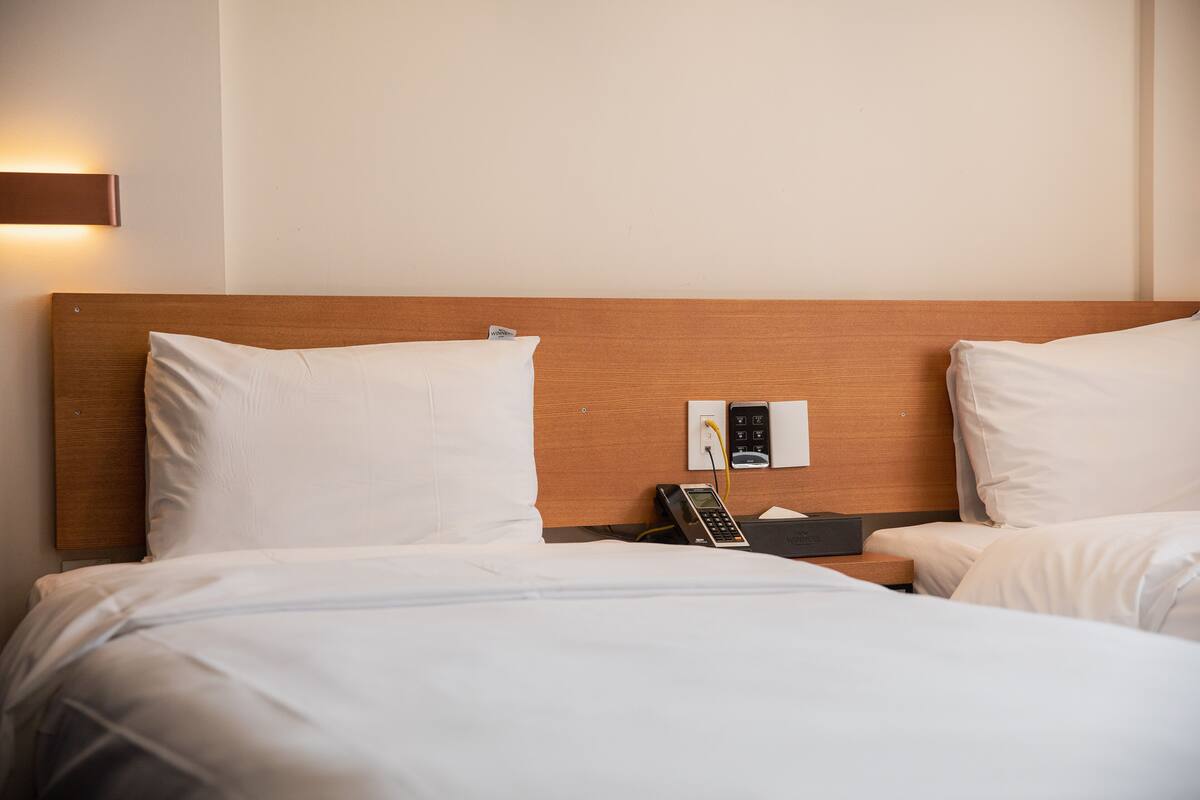 A hotel room with a wooden headboard, white bedding, a bedside table with a phone, and a wall-mounted light fixture.