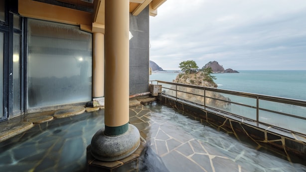 A spa area with a stone floor, a large pillar, and a view of the sea and rocky outcrops. - Hotel Yaotome (Tsuruoka)
