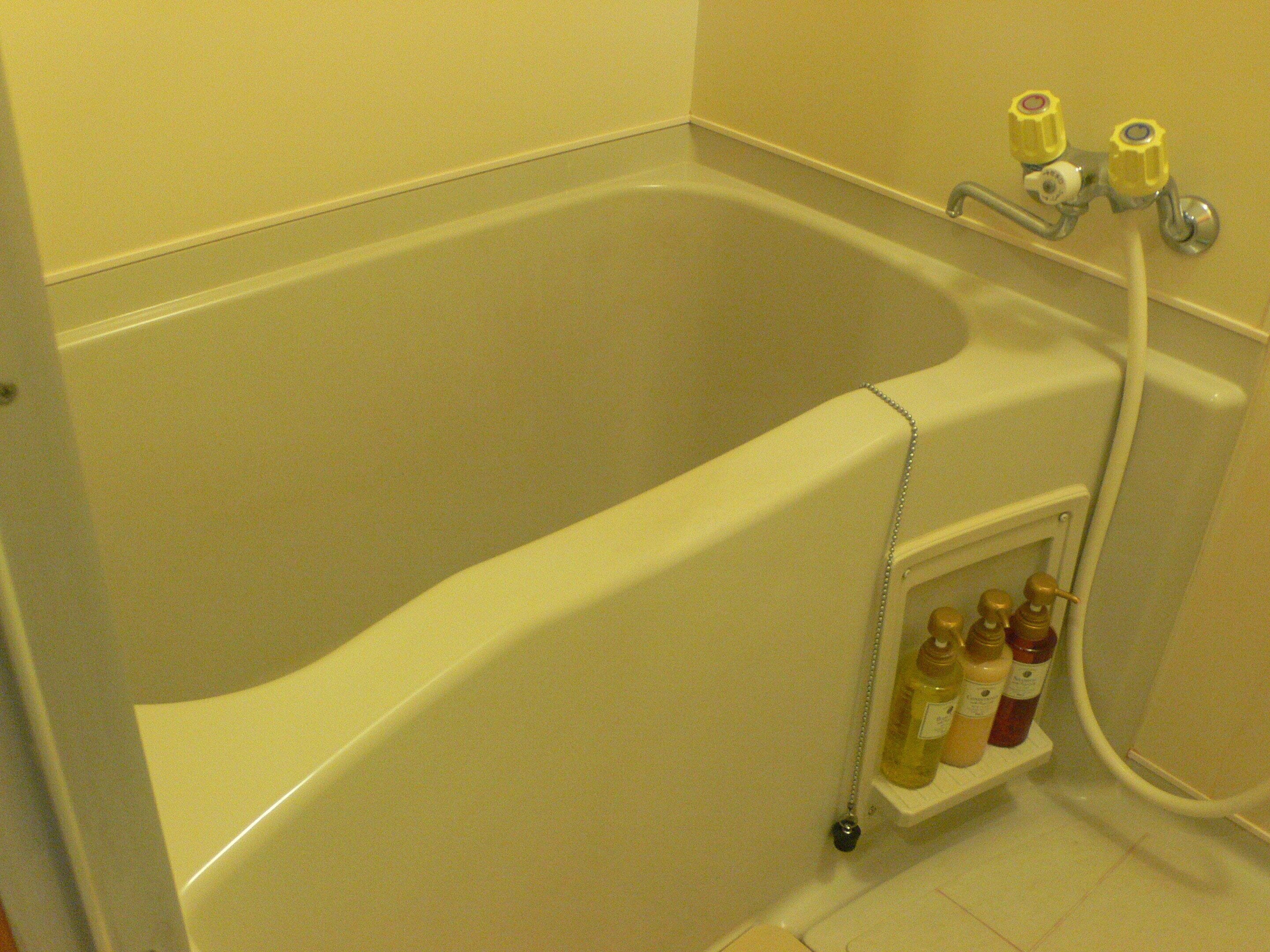 Una bañera con ducha, grifo empotrado en la pared y repisa con artículos de aseo.