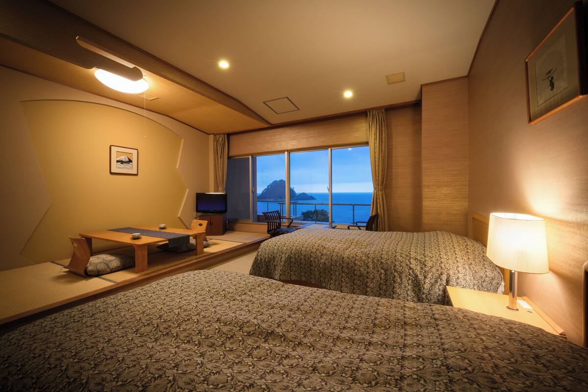 A hotel room with two beds, a desk, a chair, a TV, and a large window with a view of the sea.