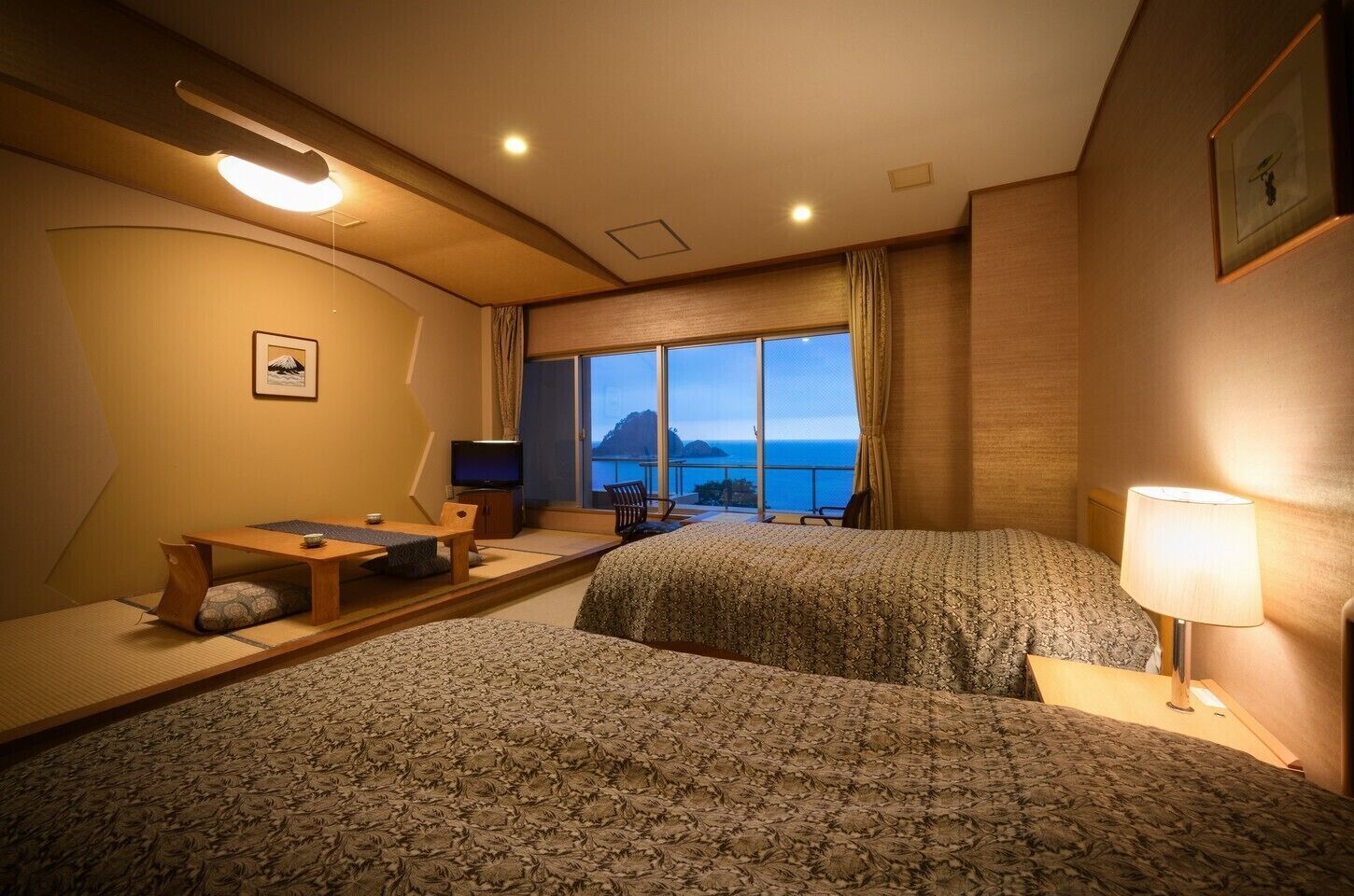 A hotel room with two beds, a desk with a chair, a television, and a view of the ocean.