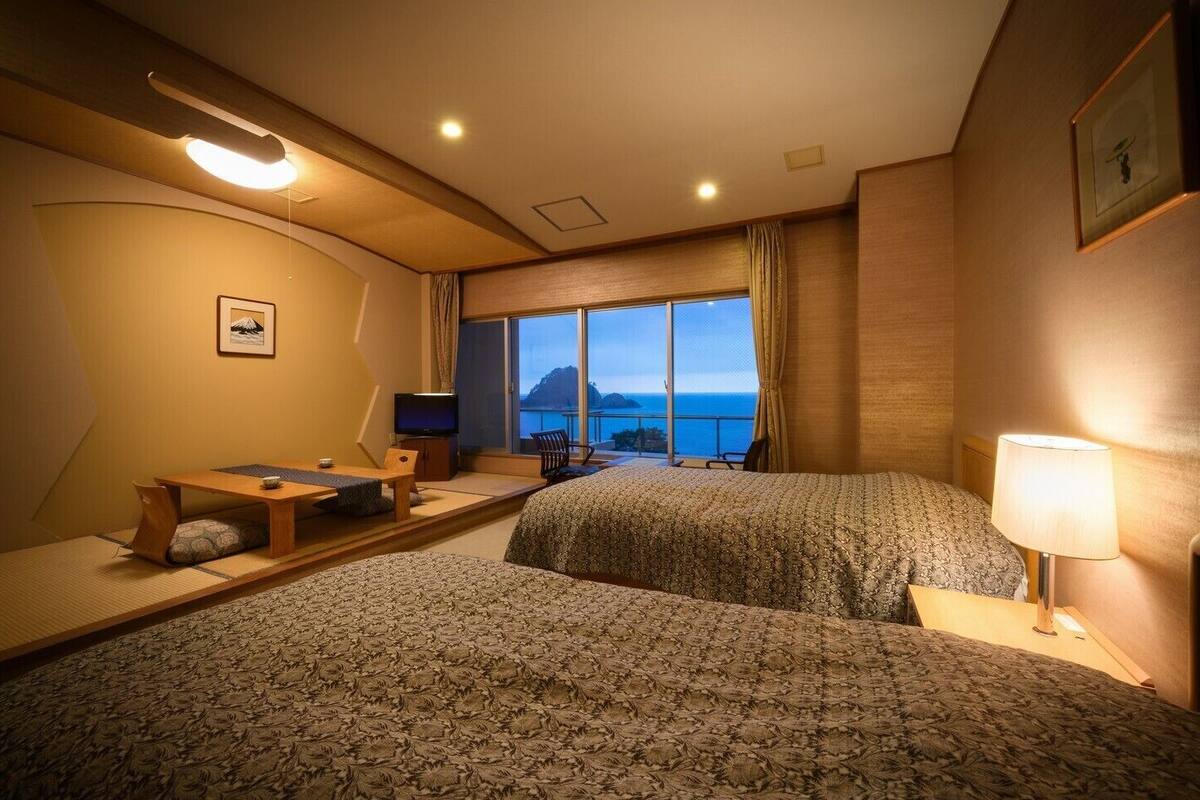 A hotel room with two beds, a desk with a chair, a television, and a view of the ocean.