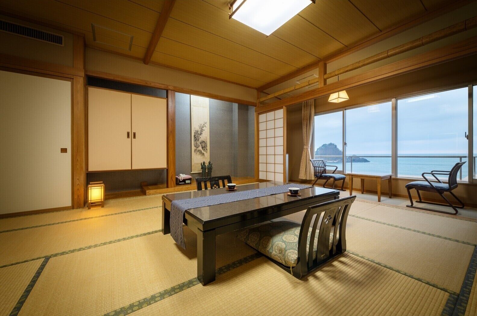A traditional Japanese room with tatami flooring, a low table with chairs, a sliding door, and a view of the sea.