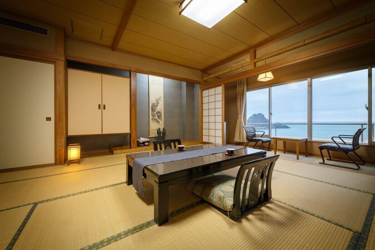 A traditional Japanese room with tatami flooring, a low table with chairs, a sliding door, and a view of the sea.