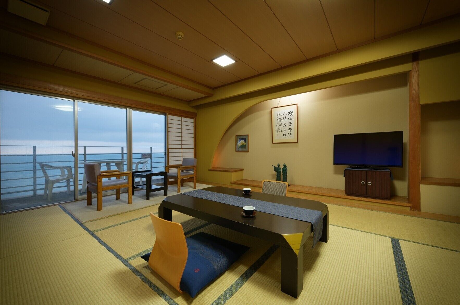 A room with tatami flooring, a low table with cushions, a television, and a view of the sea.