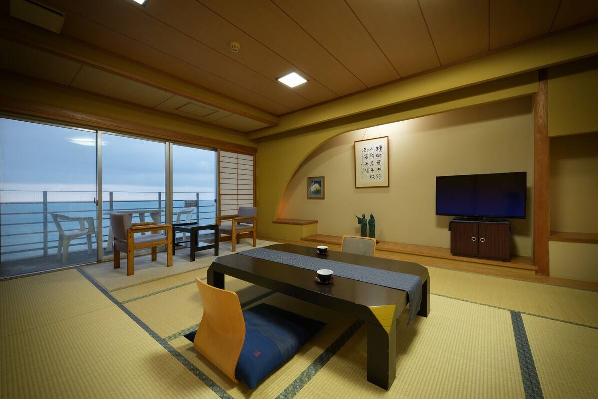 A room with tatami flooring, a television, a table, and chairs.