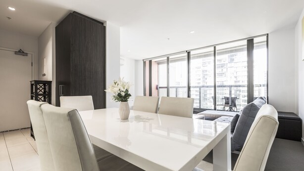 A modern living room with a white dining table, beige chairs, a dark accent wall, and a large window with a city view. - C3CBD Melbourne Apartment (Melbourne)