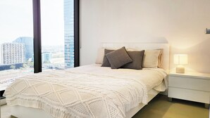A modern bedroom with a large bed, a nightstand, a lamp, and a view of a cityscape through large windows. - C3CBD Melbourne Apartment (Melbourne)