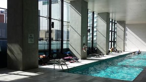 An indoor swimming pool with clear glass walls, tiled floor, and a few people swimming. - C3CBD Melbourne Apartment (Melbourne)