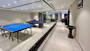 A room with two ping pong tables, a billiards table, and a glass partition.