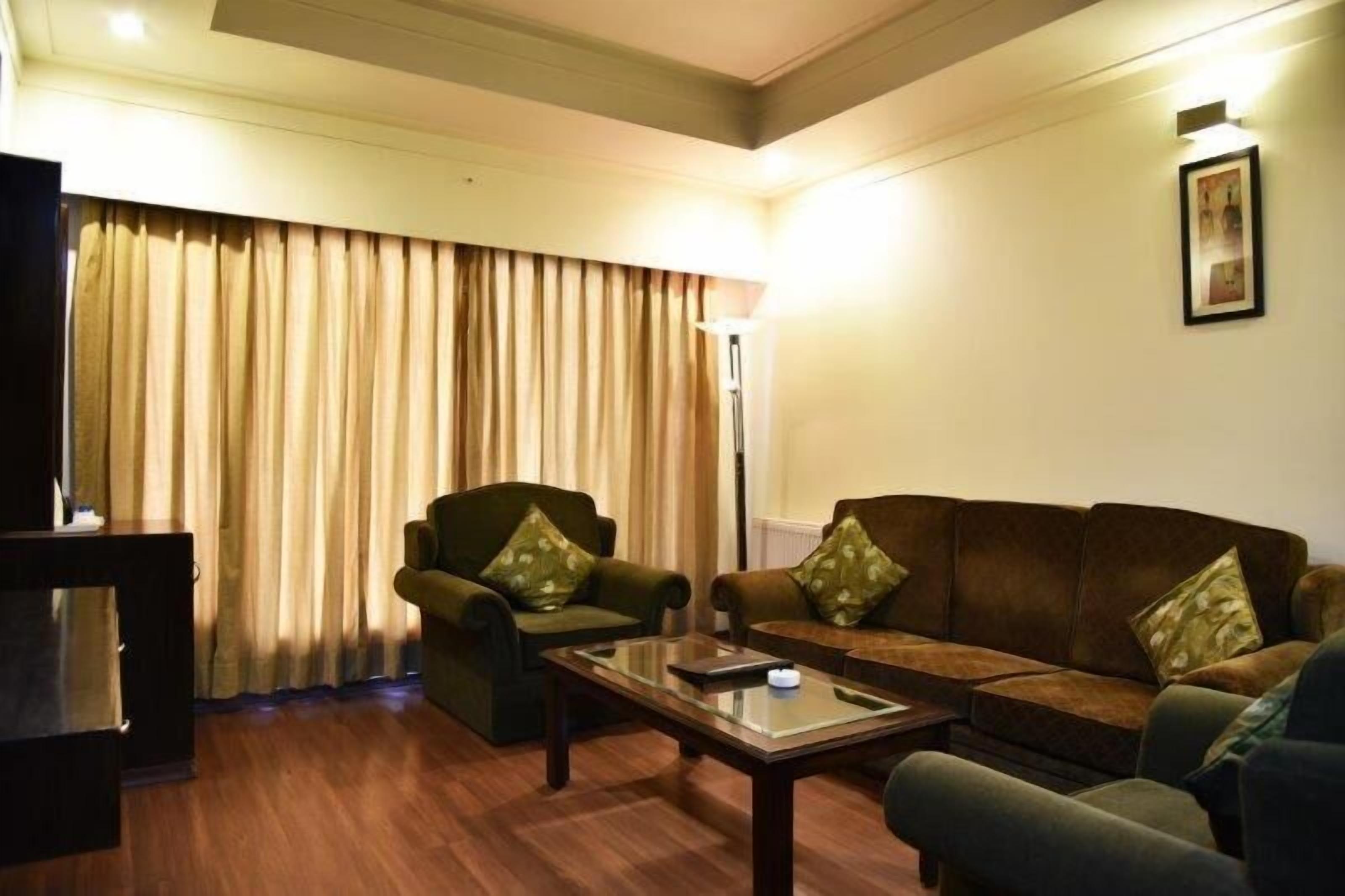 Sala de estar con un sofá, un sillón y una mesa de centro. Hay una lámpara en la pared y una cortina cubriendo una ventana.