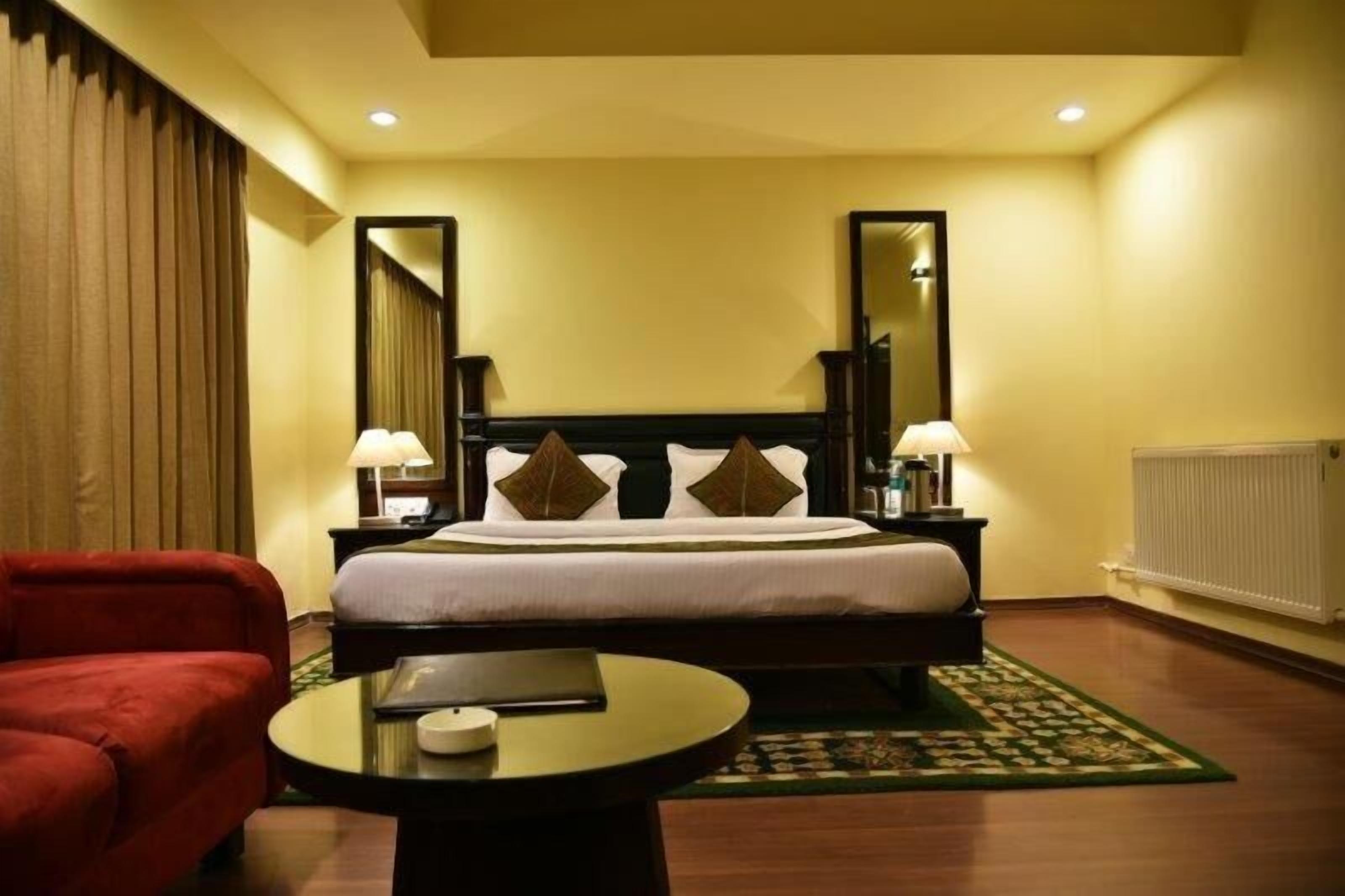 A hotel room with a bed, two bedside tables with lamps, a red armchair, a round table with a book, and a patterned rug.