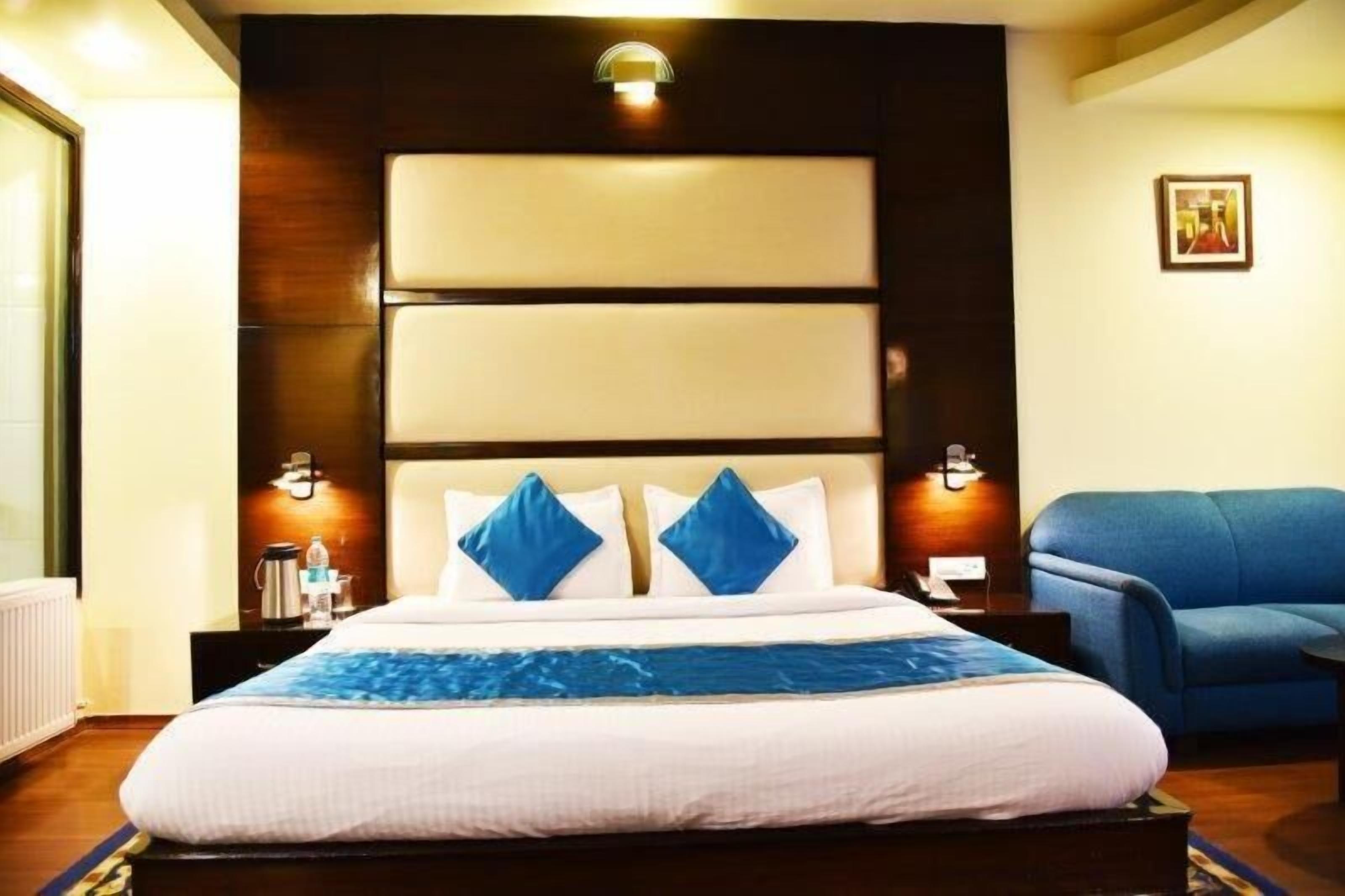 A hotel room with a large bed, two pillows, a blue throw blanket, a blue sofa, and a wall-mounted light fixture.