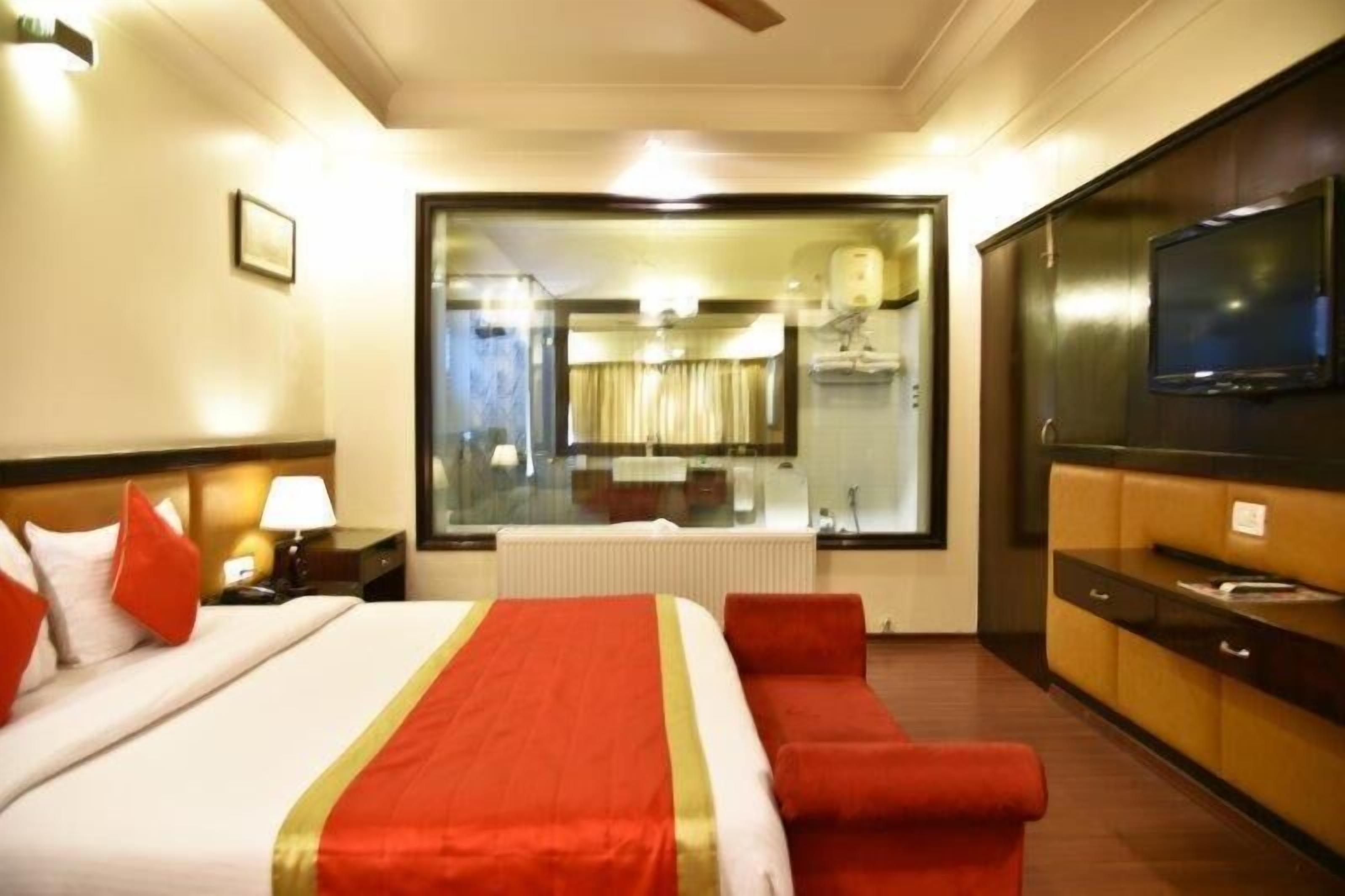 Habitación de hotel con una cama grande, televisión, escritorio y baño visible a través de un panel de vidrio.