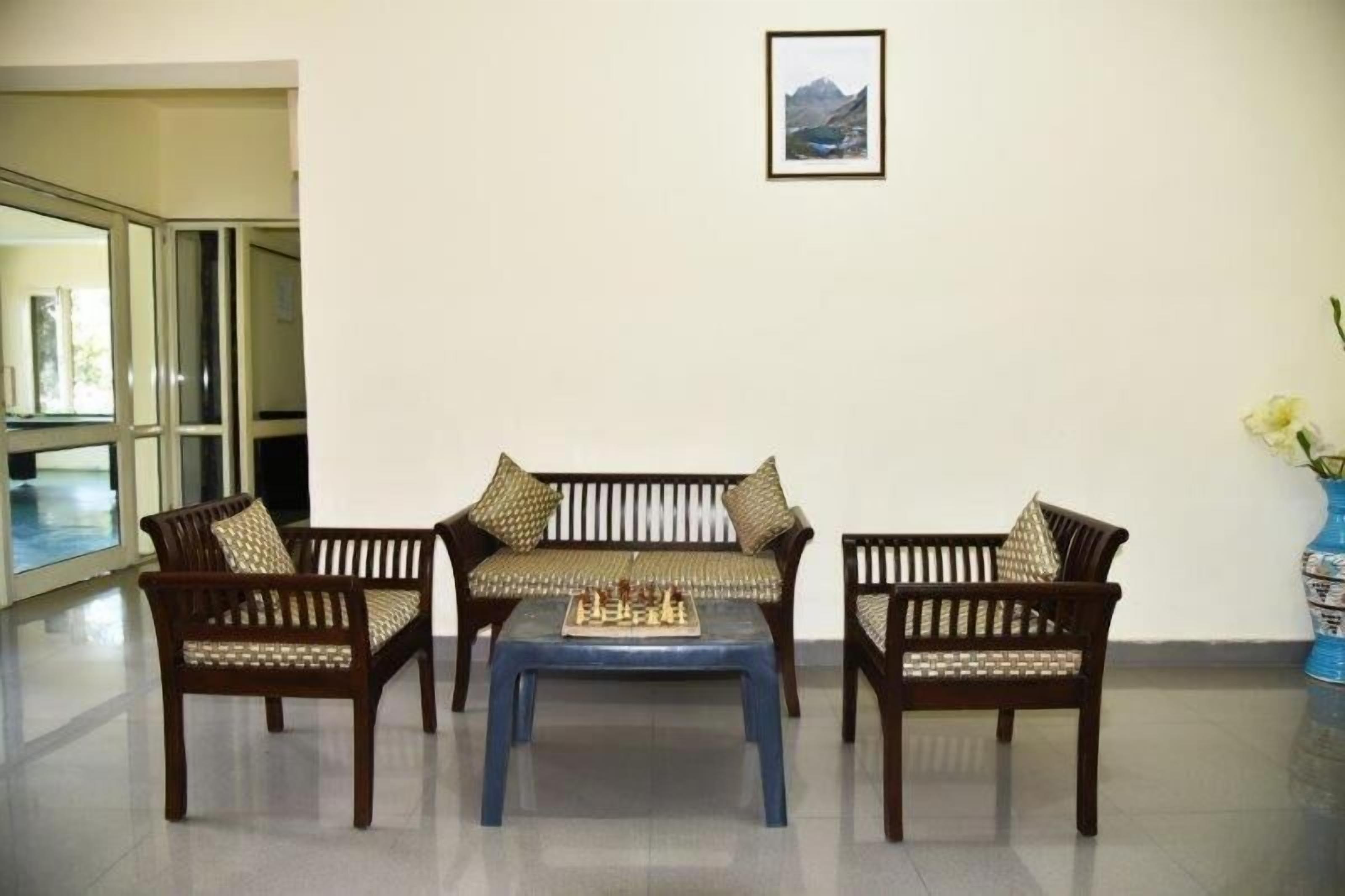 A room with three wooden chairs and a matching sofa, a chessboard on the sofa, and a framed picture on the wall.