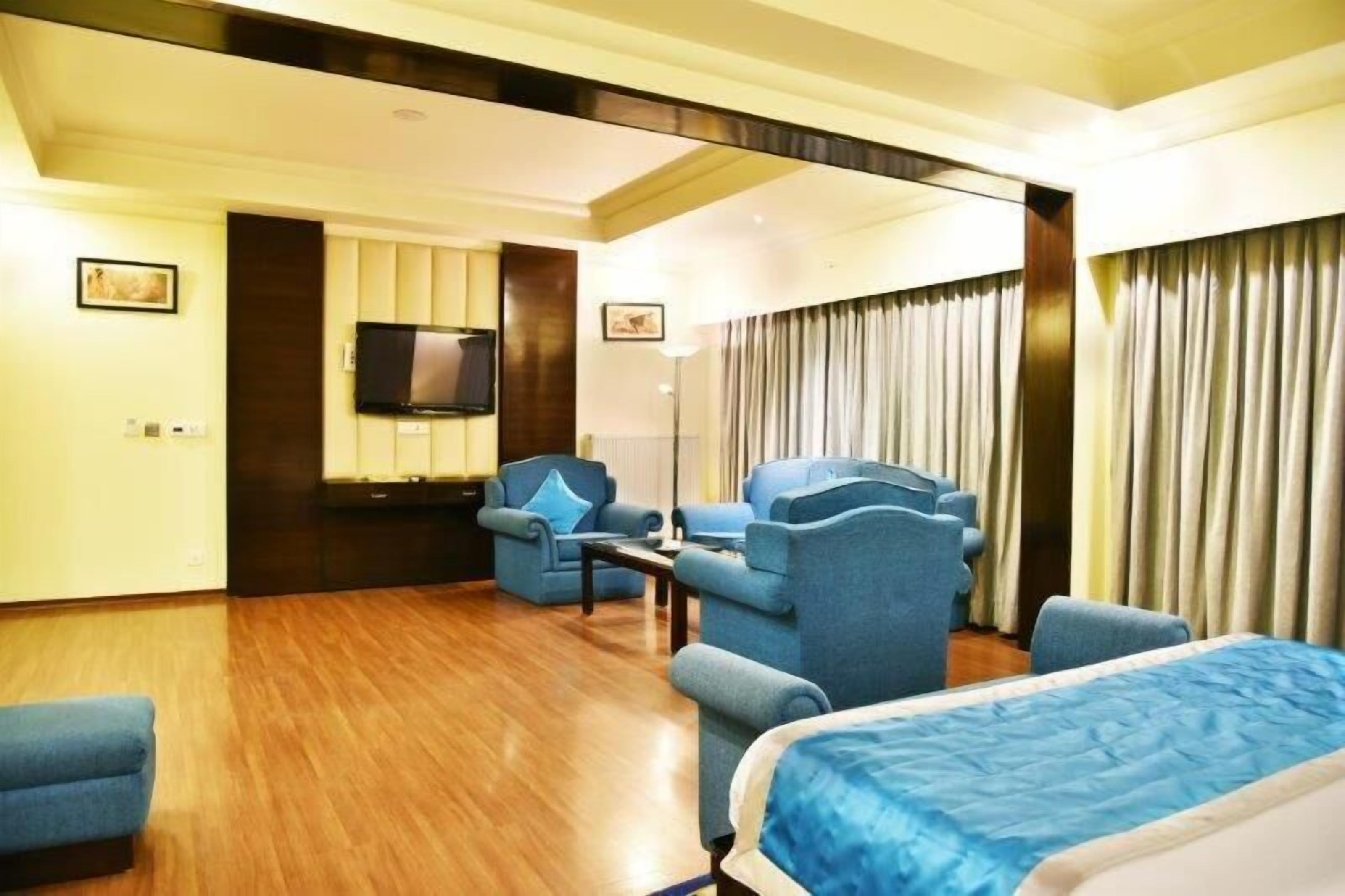 Habitación de hotel con una cama grande, una zona de estar con sillones azules, un televisor de pantalla plana montado en la pared y un ventanal con cortinas.