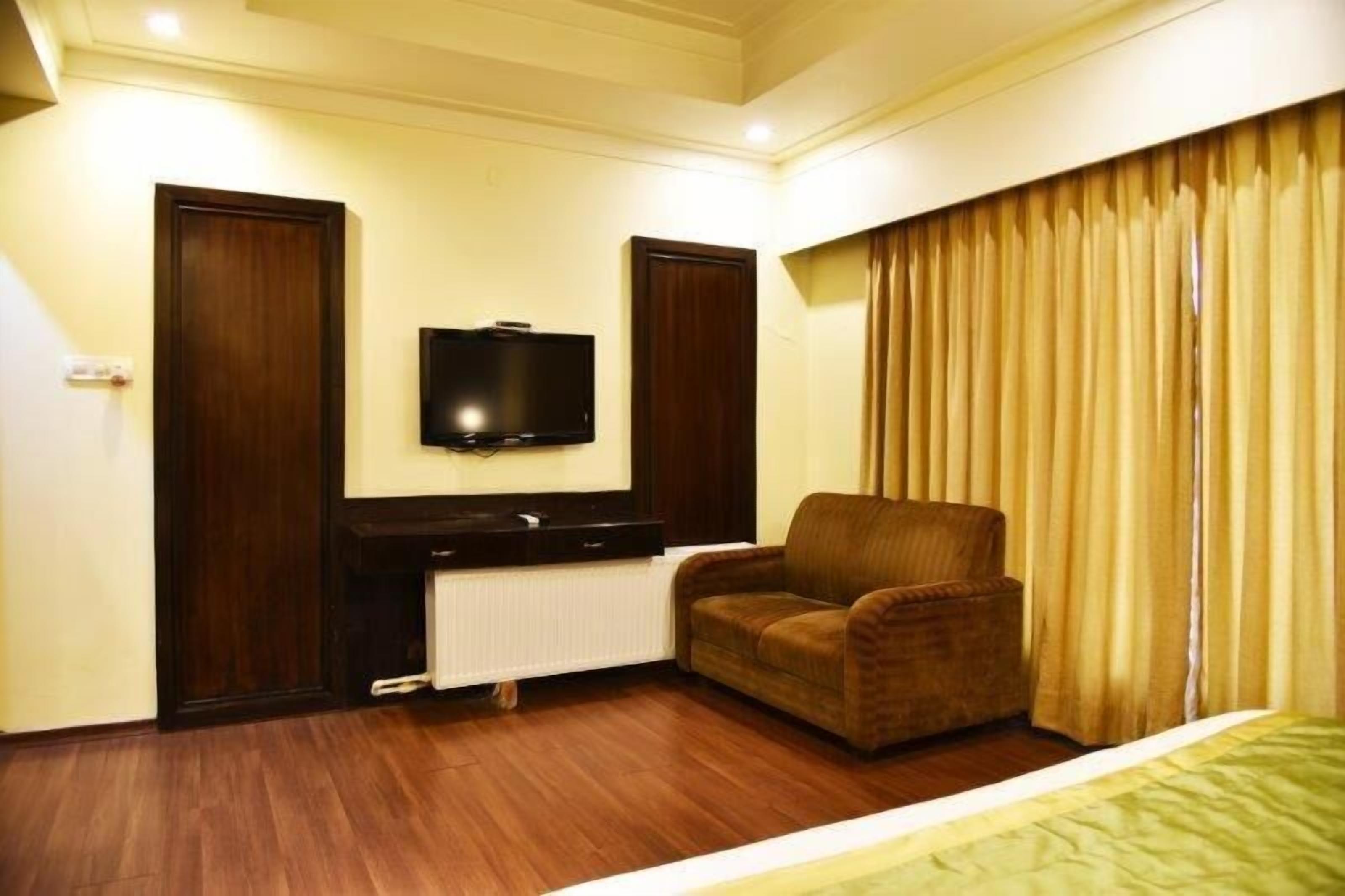 A hotel room with a wooden floor, a brown armchair, a flat-screen TV mounted on the wall, and a bed with a green bedspread.