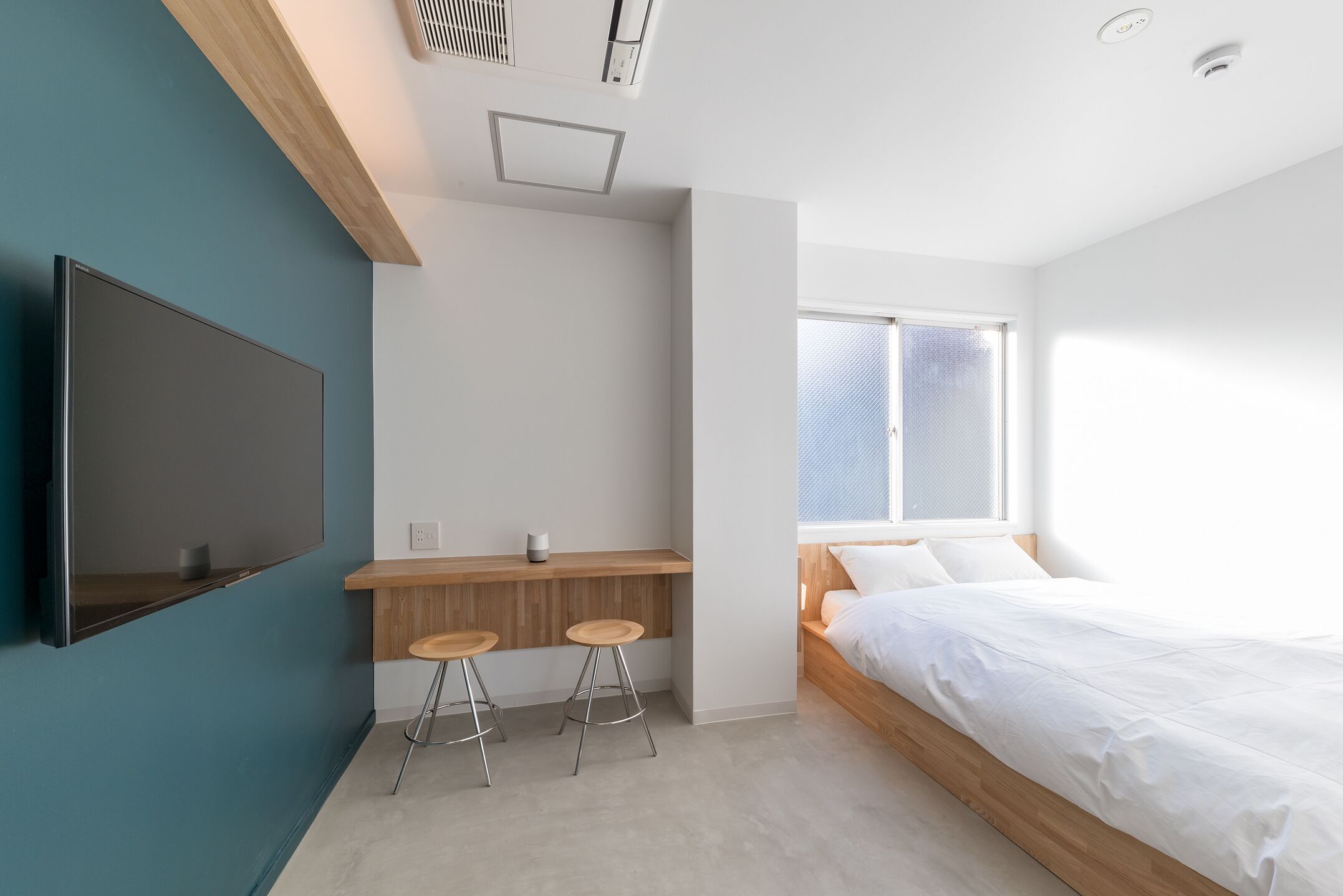 A modern bedroom with a bed, a TV mounted on the wall, a wooden desk with stools, and a window with blinds.