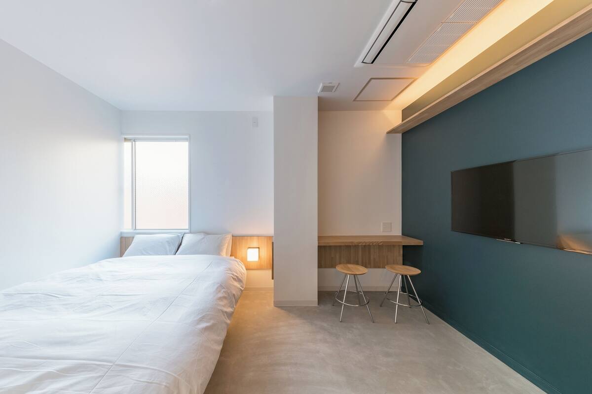 A modern bedroom with a bed, a desk, two stools, a TV, and a window.