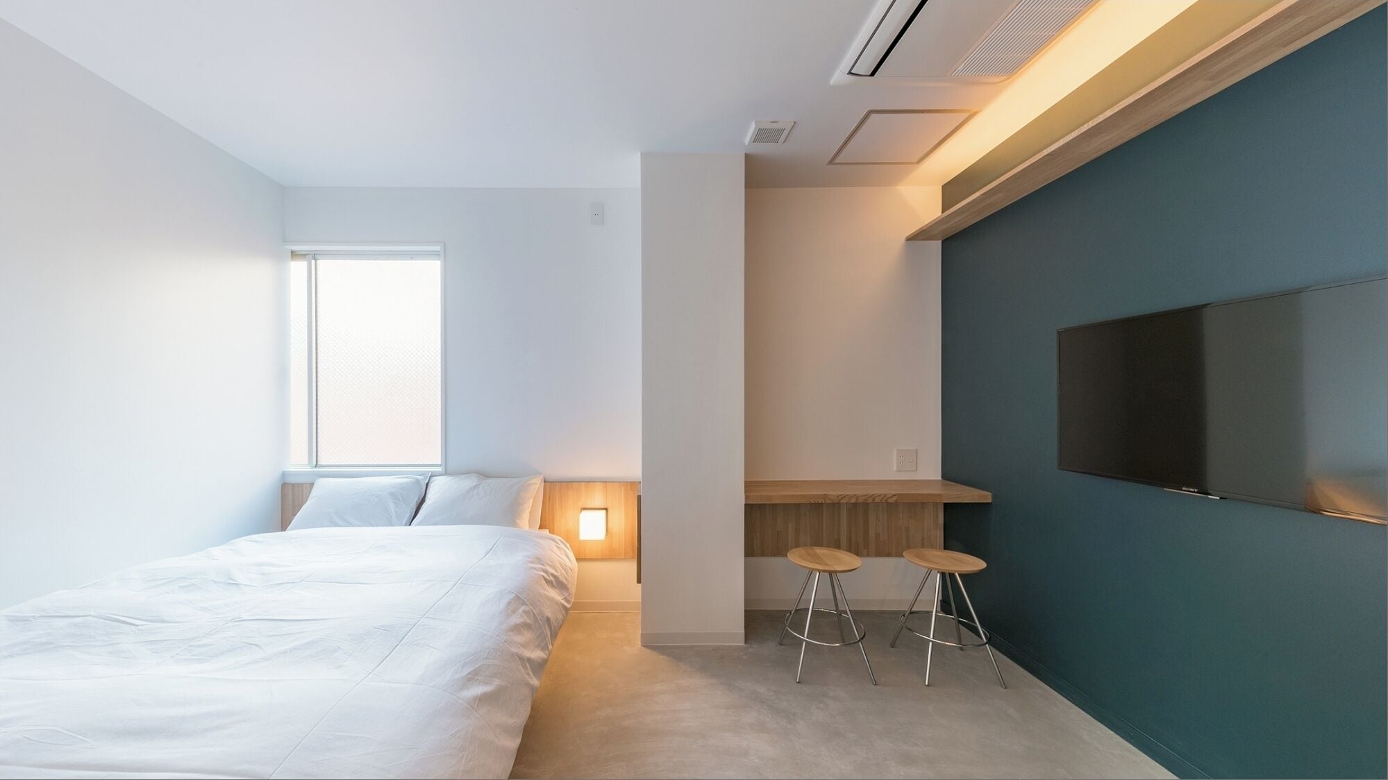 A modern hotel room with a bed, a desk with two stools, a TV mounted on the wall, and a window providing natural light.