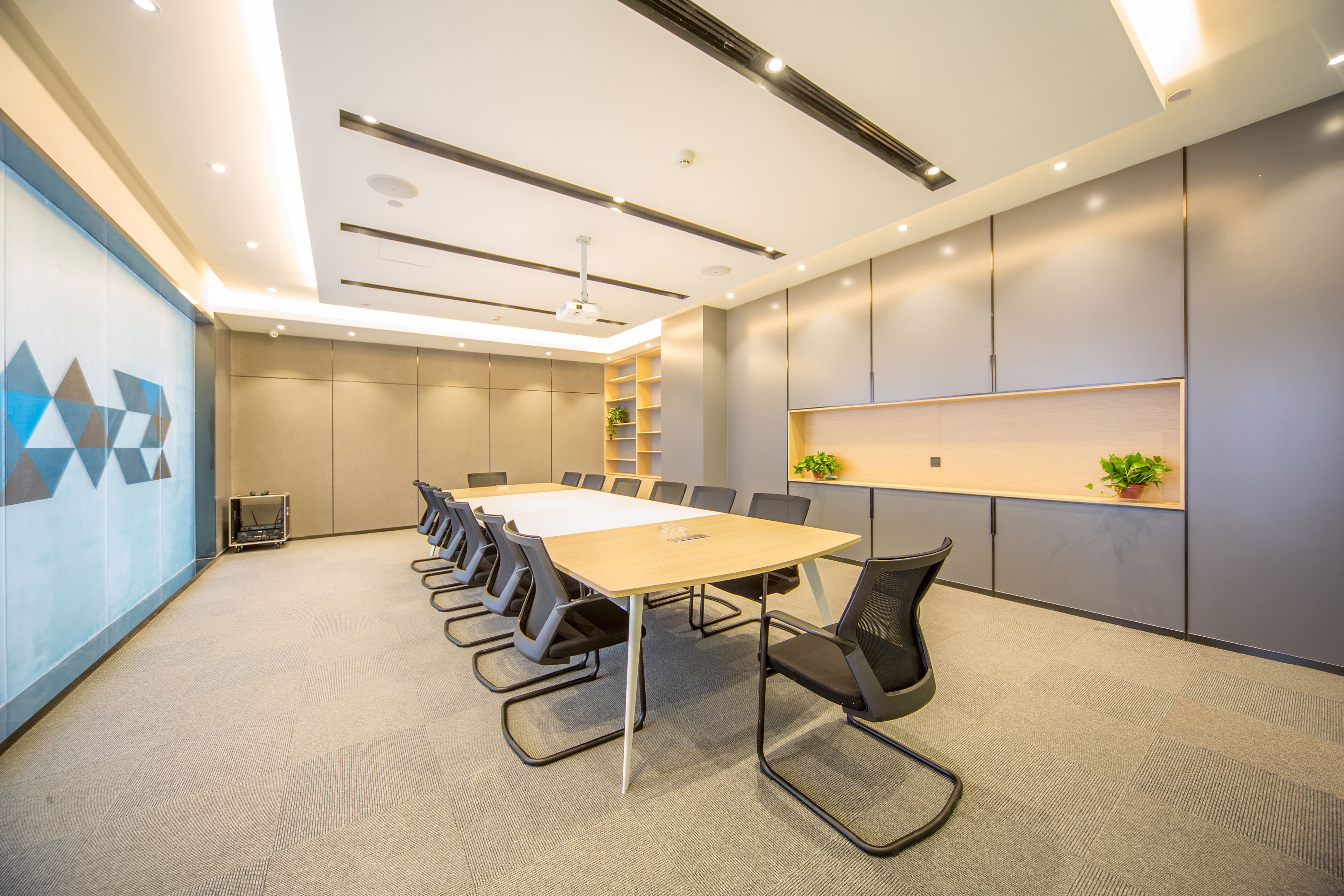 A modern conference room with a long table, black chairs, recessed lighting, and a grey carpet.