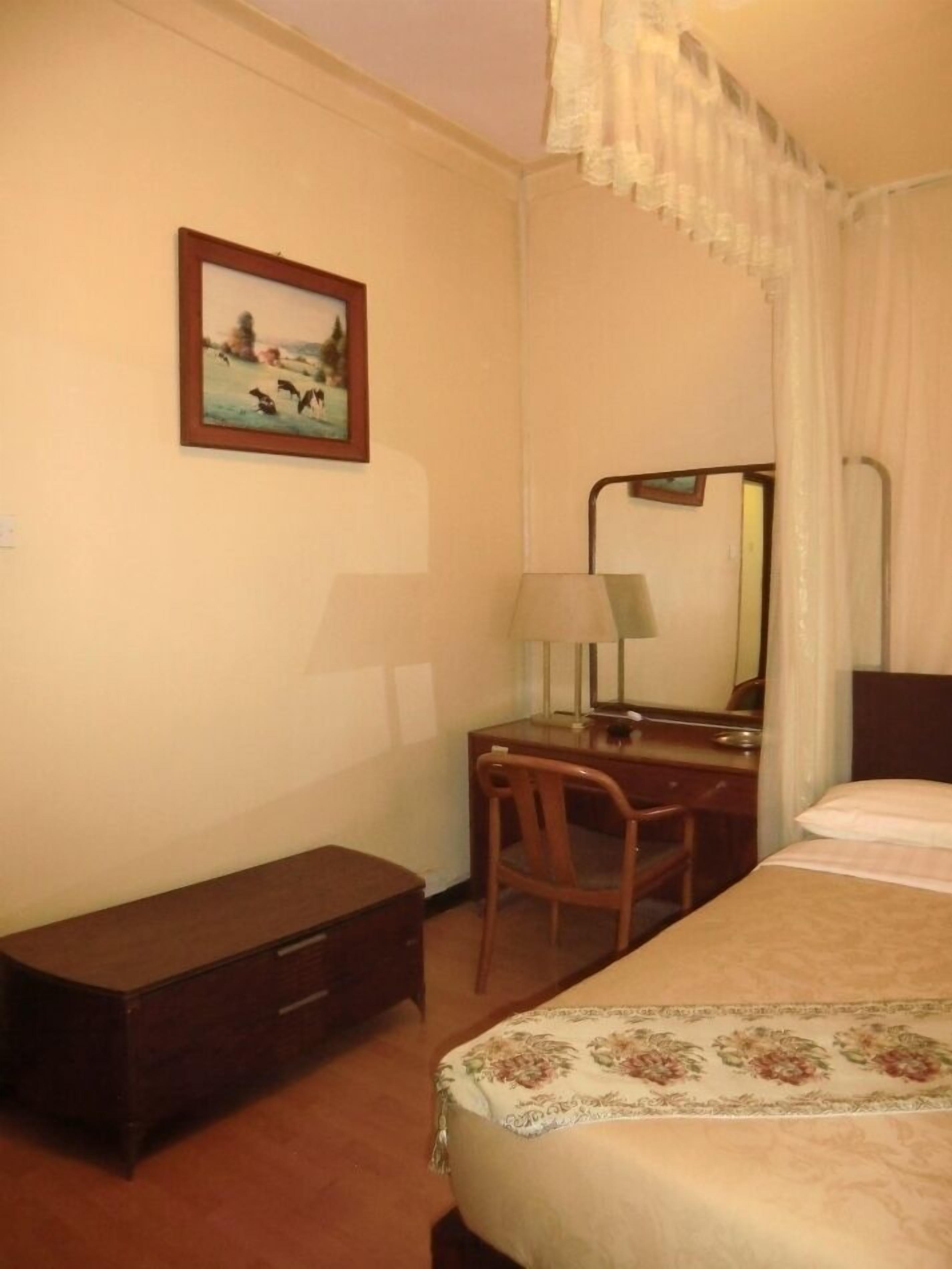 Une chambre d’hôtel avec un lit, un bureau, une chaise, une petite armoire, une lampe et un tableau au mur.