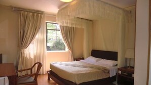 A bedroom with a four-poster canopy bed, a wooden chair, a bedside table, and a window with curtains. - Fang Fang Hotel (Kampala)