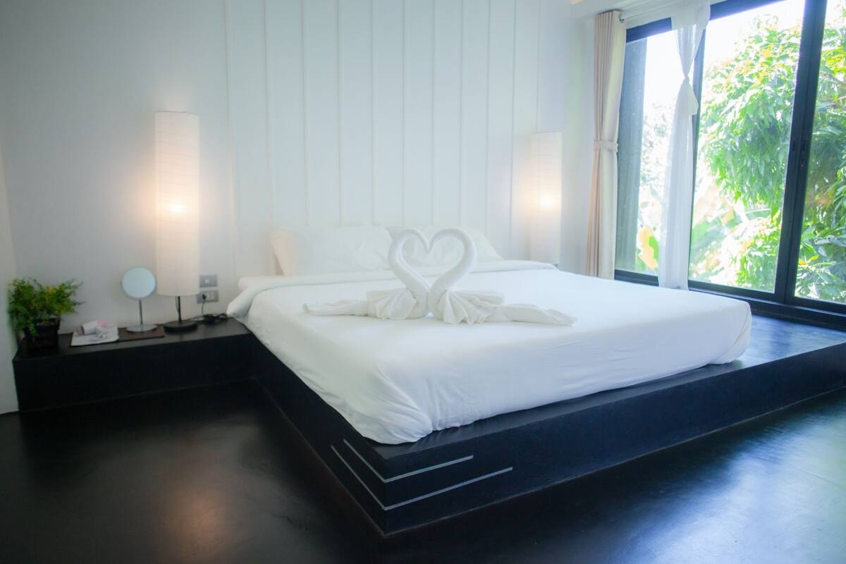 A modern hotel room with a large bed, a nightstand with a lamp, and a view of greenery through the window.