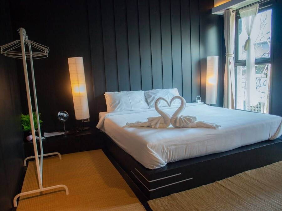 Cama bem arrumada com lençóis brancos e duas toalhas em forma de cisne, um abajur, e uma mesa de cabeceira com uma pequena planta e um objeto decorativo.